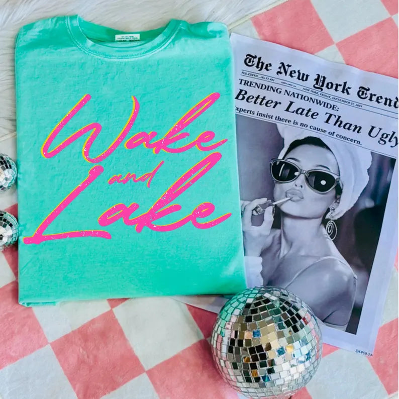 Turquoise t-shirt with pink text 'Wake and Lake' on a checkered surface with a newspaper and disco ball.