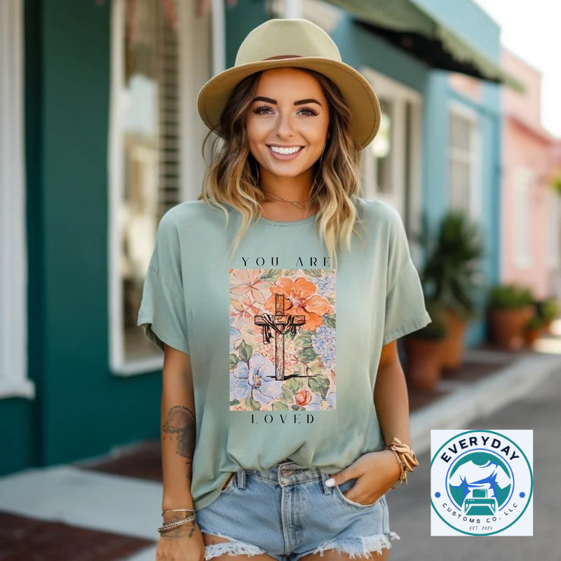 A woman in a teal t-shirt featuring a floral design and text stands outdoors.