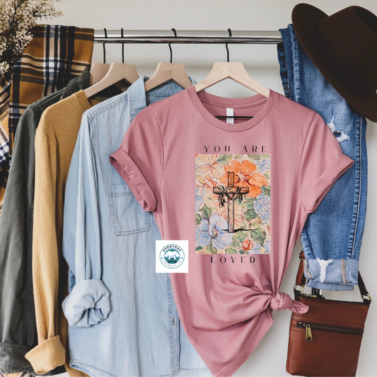 Pink t-shirt with floral design and text 'You are loved' on a rack of clothes.