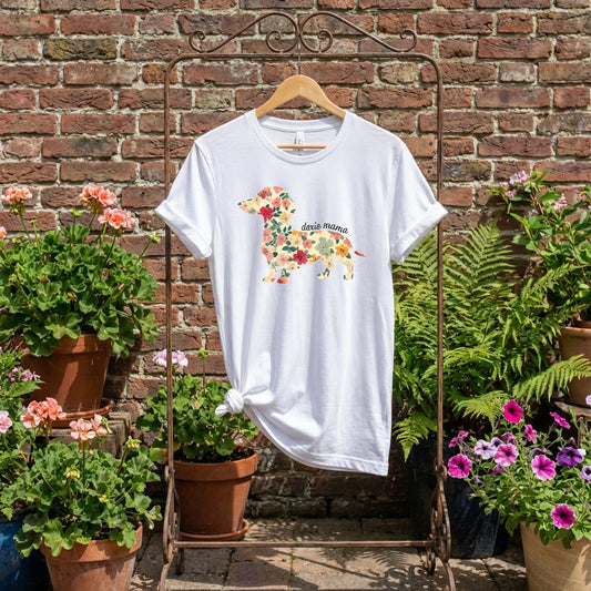 White Doxie Mama floral dachshund graphic tee hanging on a rack in a garden with brick wall and potted flowers.