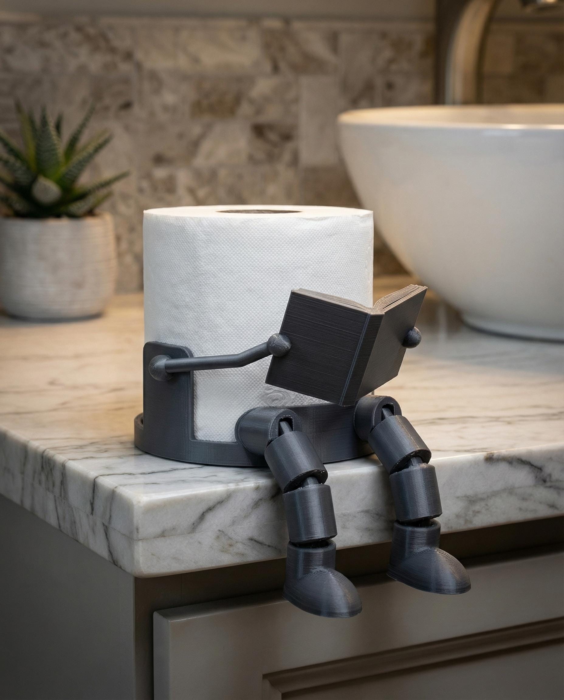 3D-printed articulated reading-man robot toilet paper holder holding a book, sits on a bathroom counter.