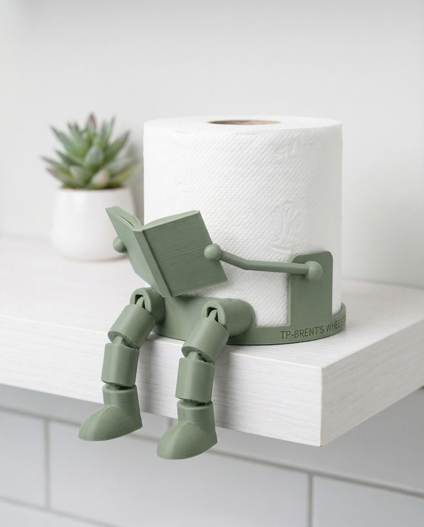 3D printed robot toilet paper holder with articulated reading man design beside a roll for bathroom decor.