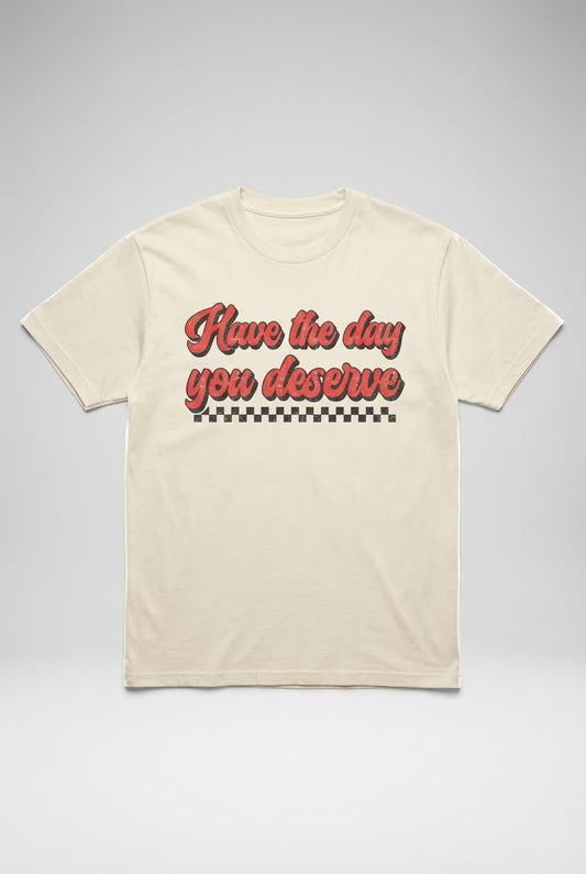 Cream graphic tee featuring retro red 'Have the day you deserve' script and checkerboard design on a studio background.