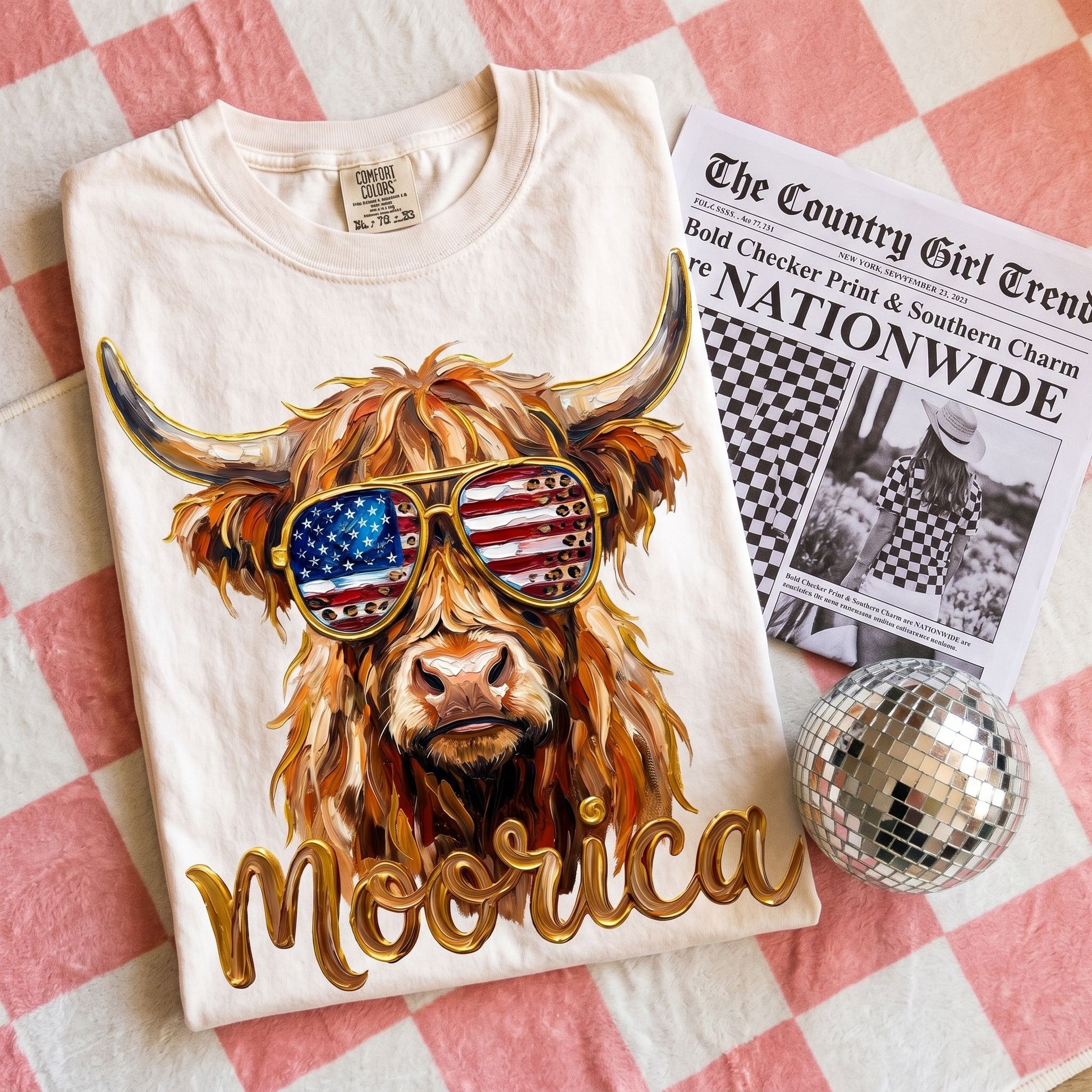 Unisex oversized Highland Cow graphic tee with checkerprint and patriotic sunglasses displayed in a warm, inviting home setting.