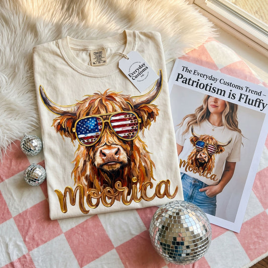 Comfort Colors Highland Cow graphic tee with American flag sunglasses, showcased in a cozy, natural living room environment.