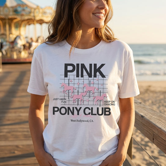 Woman wearing the Pink Pony Club white cotton graphic tee at a beach boardwalk with a carousel in the background.