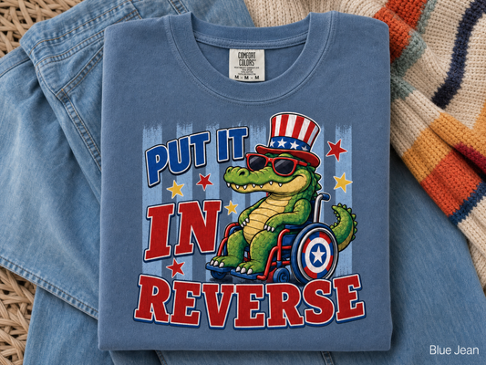 Blue t-shirt with a graphic of a crocodile in a wheelchair, sunglasses, and an American flag hat with text 'Put it in Reverse' on a blue fabric background.