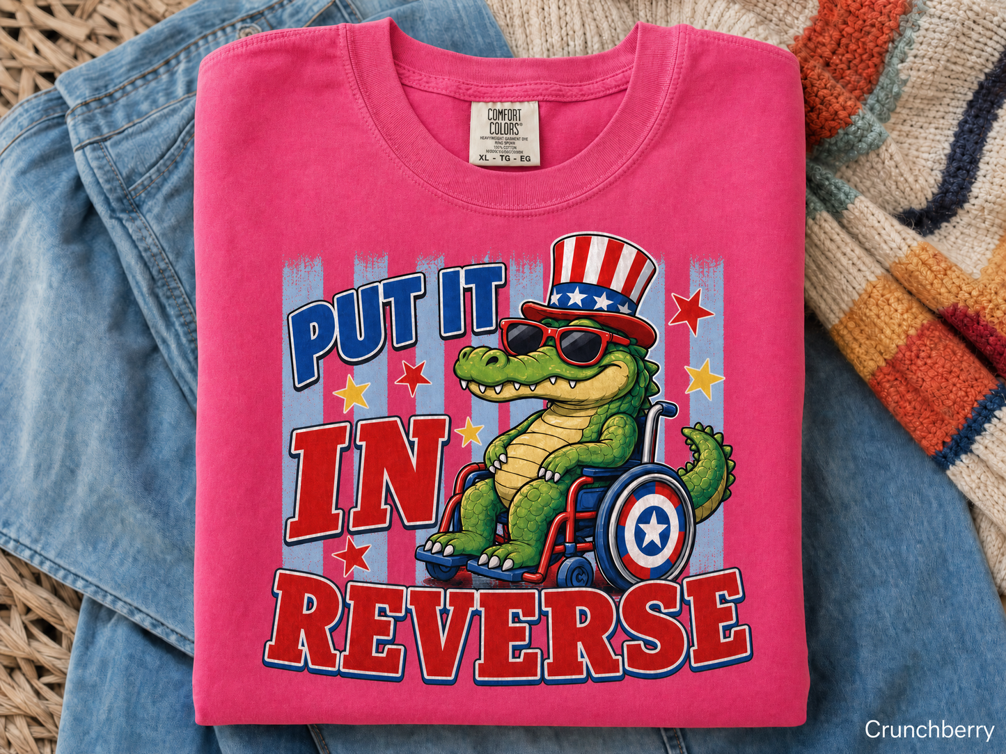 Pink t-shirt with a graphic of a crocodile in a wheelchair wearing an American flag hat, text 'Put it in reverse', on a textured surface.