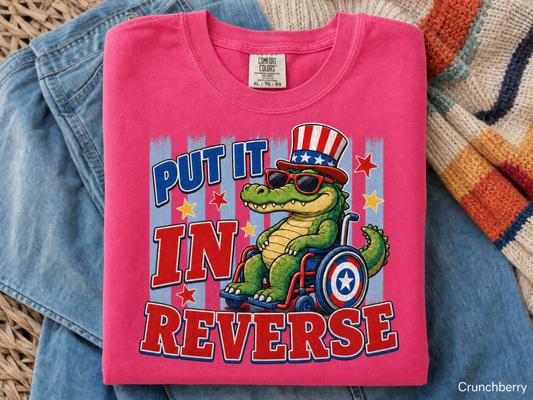 Pink t-shirt with a graphic of a crocodile in a wheelchair wearing an American flag hat, text 'Put it in reverse', on a textured surface.