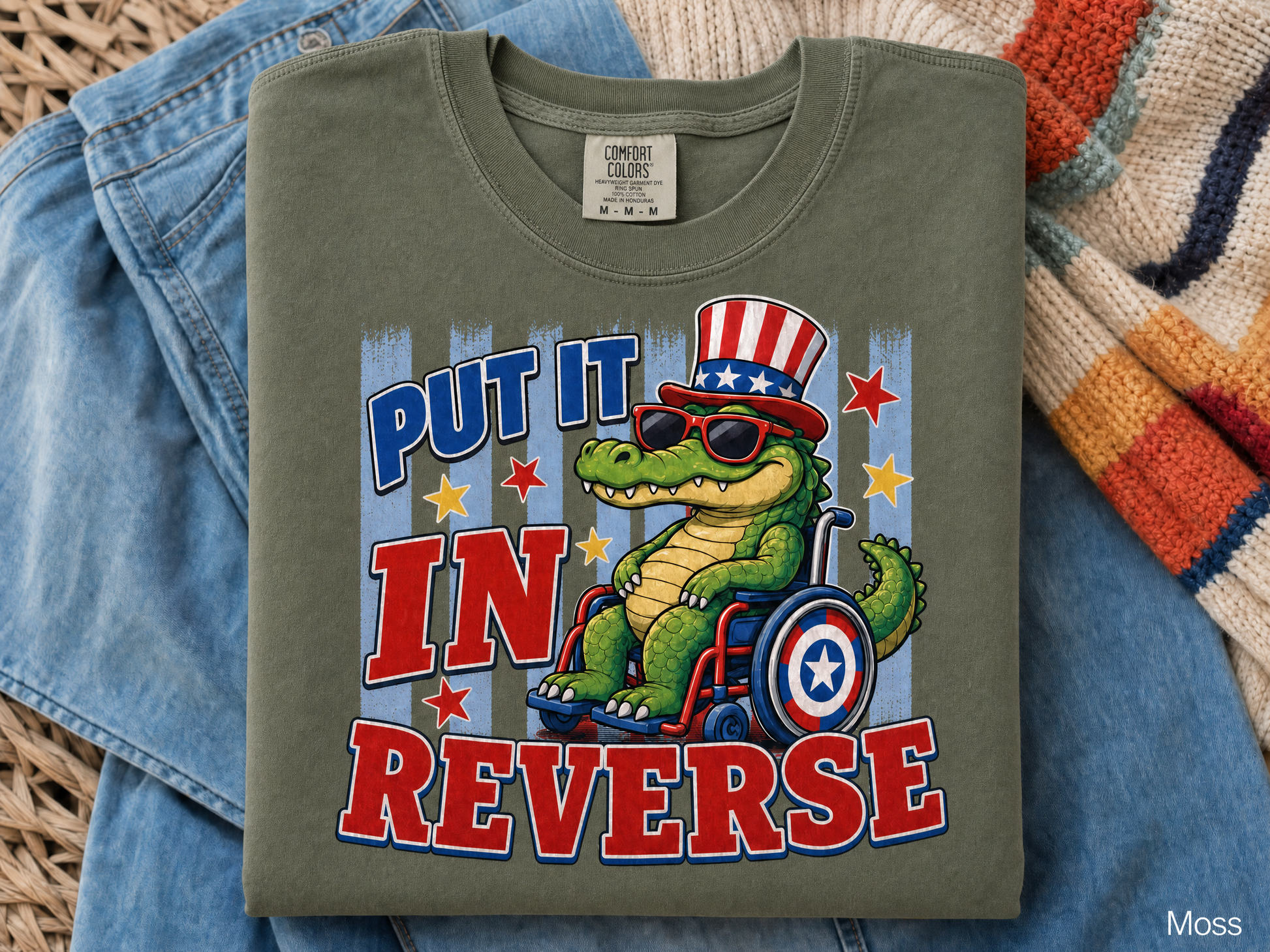Green t-shirt with a graphic of a crocodile in a wheelchair and text 'Put it in reverse' on a blue denim background.