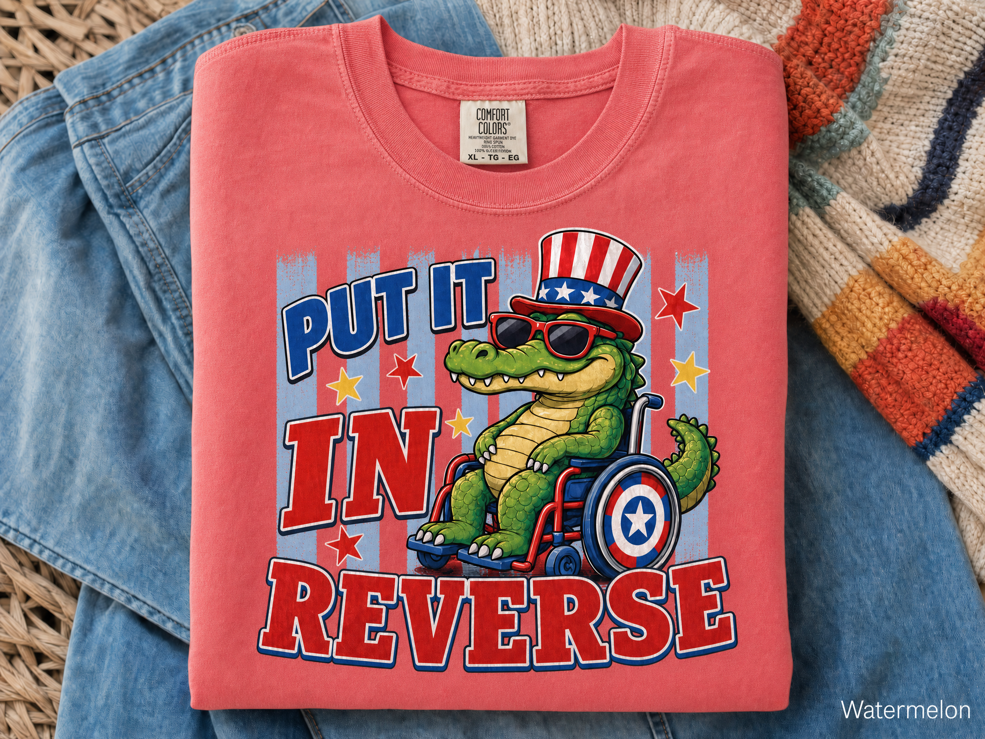 Pink t-shirt with a graphic of a crocodile in a wheelchair wearing an American flag hat, text 'Put it in Reverse', on a textured surface.